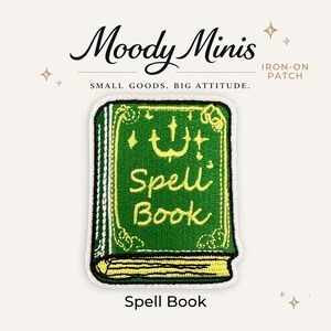 May include: An embroidered iron-on patch featuring a green book with yellow stitching. The cover displays the text "Spell Book" in yellow cursive, accented by small star symbols. The patch has a white border and is labeled "Moody Minis" and "SMALL GOODS. BIG ATTITUDE." at the top.