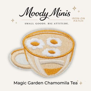 May include: An iron-on patch featuring an embroidered teacup filled with a light tan liquid and three white daisy flowers with yellow centers. The text Moody Minis, Iron-On Patch, Small Goods. Big Attitude., and Magic Garden Chamomila Tea is visible.