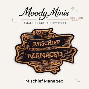May include: An iron-on patch featuring a black fabric base with gold embroidered scrollwork. The text "MISCHIEF MANAGED" is stitched in gold thread across the center of the scroll. The design includes small star accents and the brand name Moody Minis.