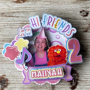 May include: A pink and purple cake topper with the words "Hi Friends" and "Maliyah" in colorful letters. The topper features a photo of a person wearing a pink shirt and a red furry puppet. The topper also includes a pink star, a blue musical note, and a pink number 2.