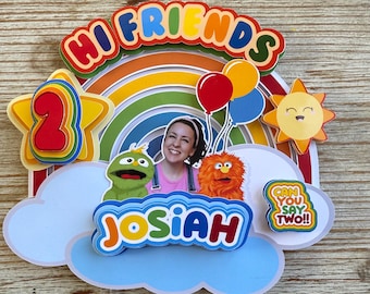 Puede incluir: Un adorno para tarta de cumpleaños colorido con las palabras "Hi Friends" y "Josiah". El diseño incluye un arcoíris, globos, un sol sonriente y personajes de dibujos animados. El número "2" se muestra de forma destacada.