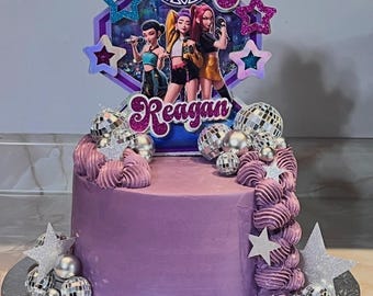 May include: A round purple frosted cake decorated with silver disco ball ornaments, glittery stars, and a custom cake topper featuring three animated characters, the number 8, and the name Reagan in purple script lettering on a hexagonal background.