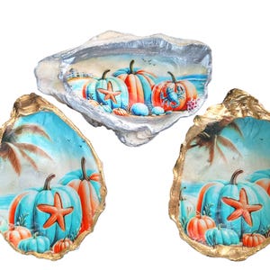 May include: Three decorative oyster shells with painted coastal scenes. Each shell features a beach landscape with pumpkins, starfish, and palm trees. The shells have silver and gold edges, and the pumpkins are teal and orange.