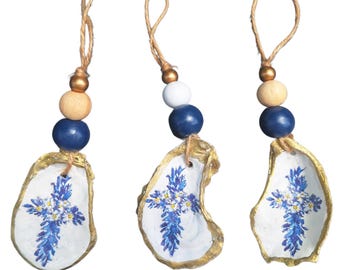 May include: Three handmade oyster shell ornaments. Each shell is painted with a blue and white cross design and trimmed with gold. They hang from a natural twine loop with wooden and blue beads.