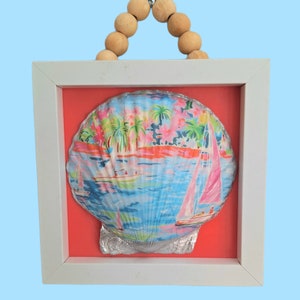 May include: A white framed seashell with a colorful print of palm trees and sailboats. The seashell is hanging from a wooden bead garland.