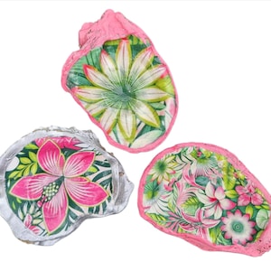 May include: Three decorative oyster shells with floral designs. The shells have pink edges and feature vibrant patterns of pink, green, and white flowers. The shells are arranged on a white background.