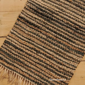 May include: A rectangular jute rug with a striped pattern in shades of brown, black, and tan. The rug has a fringed edge and is placed on a wooden surface. The rug is a natural fibre home decor item.
