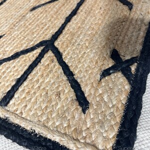 以下が含まれることがあります： 黒い幾何学模様が施された天然繊維の敷物のクローズアップ。敷物は織り込まれた質感と黒い縁取りがあります。
