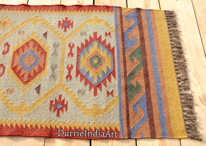 Pu&ograve; includere: Un tappeto tessuto a mano con un motivo geometrico in tonalit&agrave; di rosso, arancione, giallo, blu e verde. Il tappeto ha una frangia su un lato e il testo "DurrieIndiaArt" &egrave; visibile nell'angolo in basso a destra.