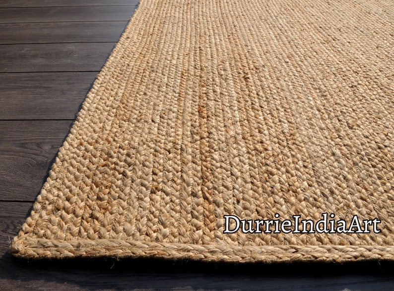 K&ouml;nnte beinhalten: Ein Naturfaser-Teppich aus Jute, einem nachhaltigen und strapazierf&auml;higen Material. Der Teppich hat eine gewebte Textur und eine nat&uuml;rliche beige Farbe. Der Teppich ist auf einem Holzboden abgebildet. Der Text "DurrieIndiaArt" ist in der unteren rechten Ecke des Bildes sichtbar.