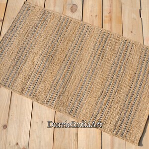 May include: Rectangular jute rug with a natural tan color and vertical stripes of dark gray. The rug is placed on a light-colored wooden surface. The rug's texture is visible, suggesting a woven construction. The rug is suitable for home decor.