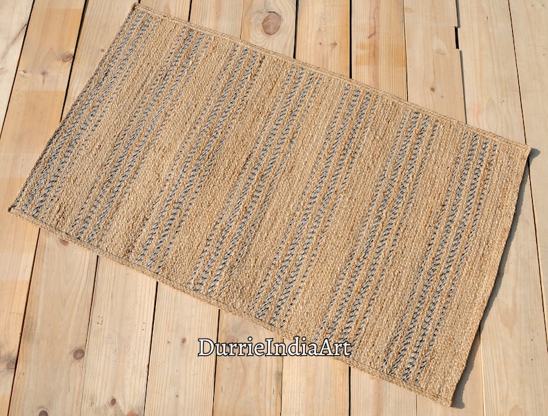May include: Rectangular jute rug with a natural tan color and vertical stripes of dark gray. The rug is placed on a light-colored wooden surface. The rug's texture is visible, suggesting a woven construction. The rug is suitable for home decor.
