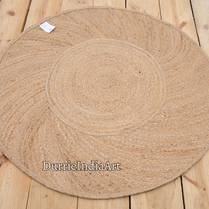 Può includere: Un tappeto rotondo in fibra naturale con un design intrecciato. Il tappeto è in juta e ha un colore beige naturale. Il tappeto ha un diametro di circa 90 cm. Il testo "DurrieIndiaArt" è stampato sul tappeto.