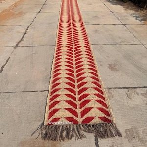 Puede incluir: Una alfombra de pasillo larga y estrecha con un patrón de espiga repetitivo en fibras de color rojo intenso y bronceado natural. La alfombra tiene una apariencia tejida y texturizada con una costura vertical central y flecos deshilachados.