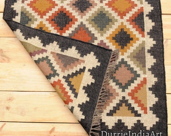 May include: A black and white rug with a geometric pattern in shades of brown, orange, and green. The rug has a fringe border and is folded in half. The text "DurrieIndiaArt" is visible in the bottom right corner.
