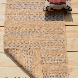 May include: A rectangular jute rug with a woven pattern of natural and gray stripes. A decorative pillow with a red, black, and cream geometric design rests on the rug. The rug is on a wooden surface.