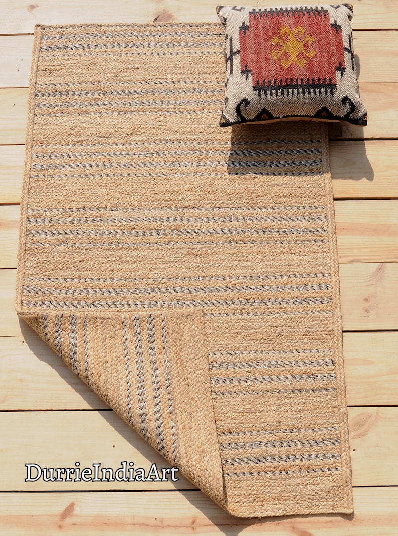 May include: A rectangular jute rug with a woven pattern of natural and gray stripes. A decorative pillow with a red, black, and cream geometric design rests on the rug. The rug is on a wooden surface.