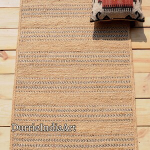 May include: A rectangular jute rug with a striped pattern in natural and gray tones. A decorative pillow with a geometric design in red, black, and cream rests on the rug. The rug is on a wooden surface.