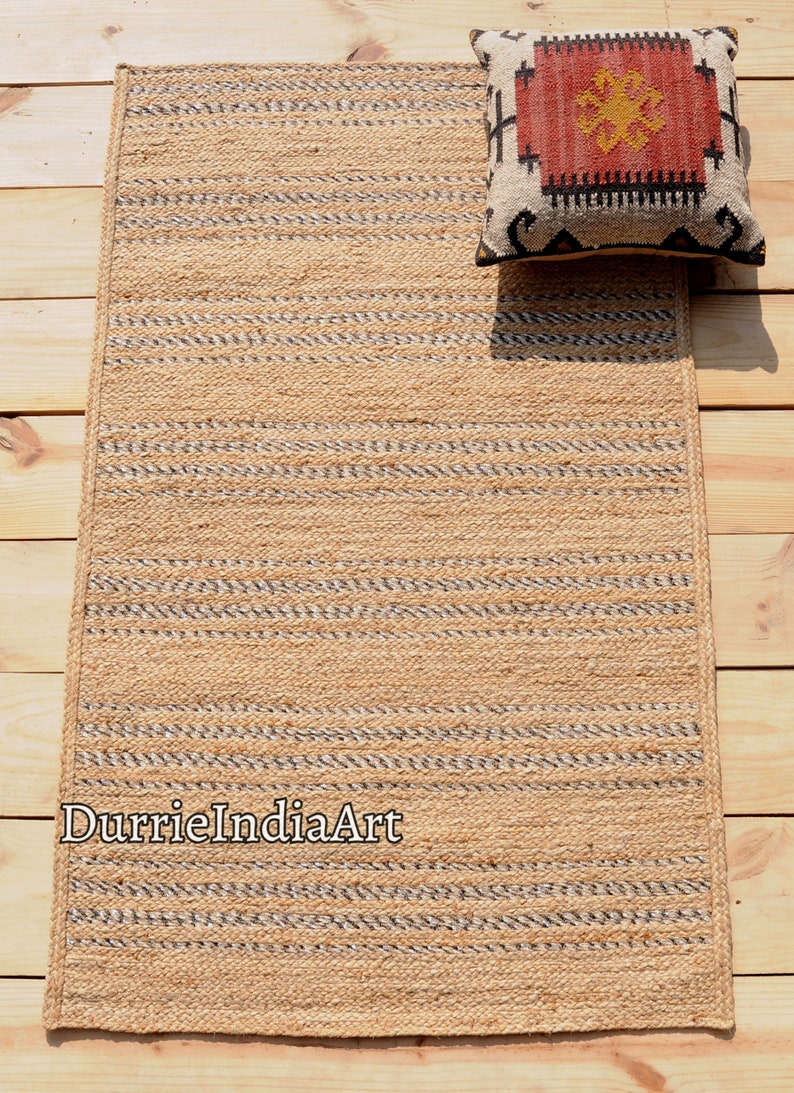 May include: A rectangular jute rug with a striped pattern in natural and gray tones. A decorative pillow with a geometric design in red, black, and cream rests on the rug. The rug is on a wooden surface.