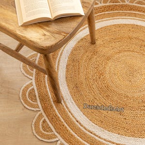 May include: A round jute rug with a white border and a scalloped edge. The rug is made from natural fibres and has a rustic, earthy look. The text "DurrieIndiaArt" is printed on the rug.