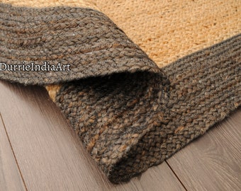 May include: Close-up of a natural fiber rug with a brown and beige color scheme. The rug has a thick, woven texture and a dark brown border. The rug is rolled up on a wooden floor.