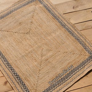 Puede incluir: Una alfombra de fibra natural con un dise&ntilde;o tejido y un borde gris oscuro. La alfombra est&aacute; hecha de yute y tiene un aspecto natural y terroso.
