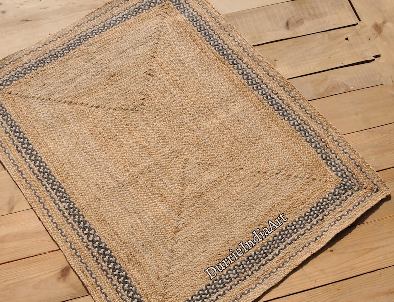 Puede incluir: Una alfombra de fibra natural con un dise&ntilde;o tejido y un borde gris oscuro. La alfombra est&aacute; hecha de yute y tiene un aspecto natural y terroso.