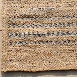 May include: Close-up of a natural jute rug with woven stripes in black and silver. The rug has a braided border and the text "DurrieIndiaArt" is visible in the upper right corner. The rug is made of a natural fiber.