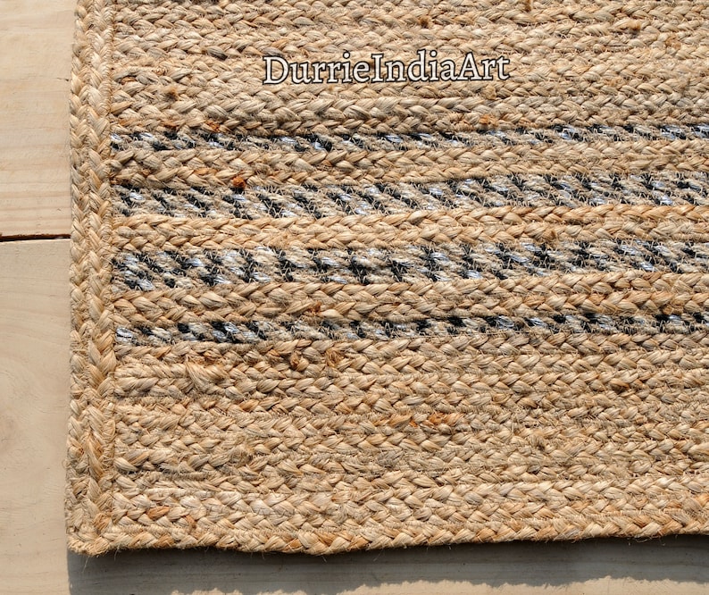 May include: Close-up of a natural jute rug with woven stripes in black and silver. The rug has a braided border and the text "DurrieIndiaArt" is visible in the upper right corner. The rug is made of a natural fiber.