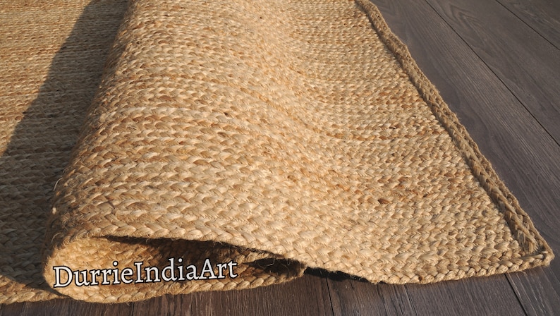 Pode incluir: Um tapete de fibra natural feito de juta, um material sustent&aacute;vel e dur&aacute;vel. O tapete tem uma textura tecida e uma cor bege natural. O tapete est&aacute; enrolado e a borda &eacute; vis&iacute;vel. O texto "DurrieIndiaArt" &eacute; vis&iacute;vel no tapete.