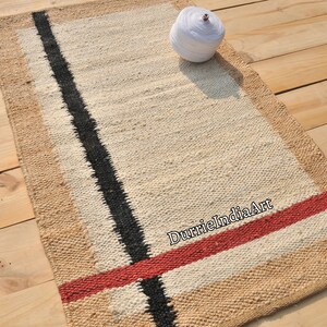 May include: A rectangular rug with a white background and black and red stripes. The rug is made of natural fibers and has a woven texture. The text "DurrieIndiaArt" is visible in the lower right corner of the rug.