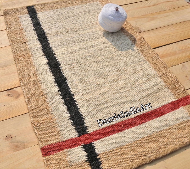 May include: A rectangular rug with a white background and black and red stripes. The rug is made of natural fibers and has a woven texture. The text "DurrieIndiaArt" is visible in the lower right corner of the rug.