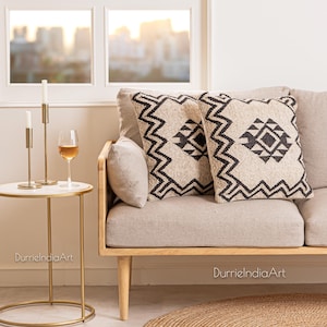 May include: Two black and white geometric patterned throw pillows on a light beige sofa with wooden legs. The sofa is in a room with a window and a side table with a glass of wine and candles.