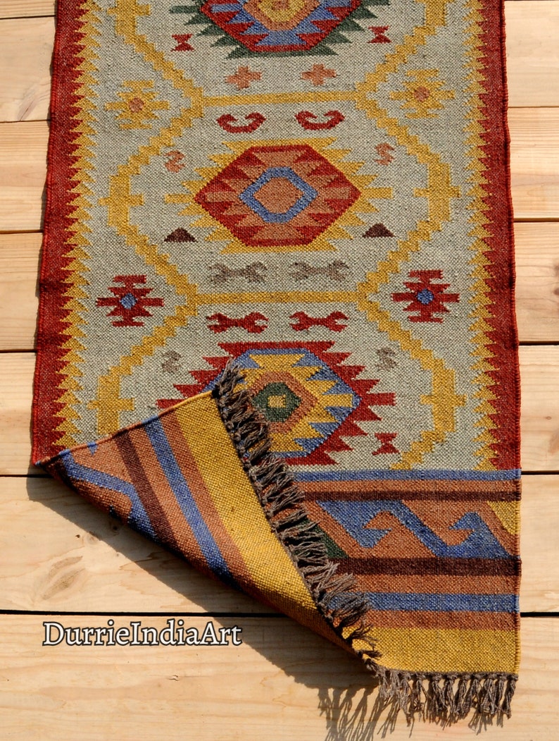 Pu&ograve; includere: Un tappeto colorato tessuto a mano con un motivo geometrico in tonalit&agrave; di rosso, giallo, blu e verde. Il tappeto ha un bordo a frangia ed &egrave; piegato a met&agrave;, rivelando il retro.