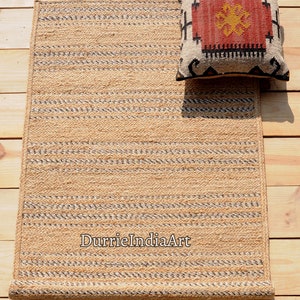 May include: A rectangular jute rug with horizontal stripes in natural and gray tones. A decorative pillow with a red, yellow, and black geometric design rests on the rug. The text "DurrieIndiaArt" is visible on the rug.