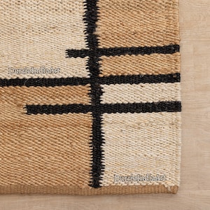 Puede incluir: Un primer plano de una alfombra de yute tejida con un patrón geométrico en negro, blanco y beige. La alfombra tiene una sensación natural y terrosa y es perfecta para agregar un toque de textura a cualquier habitación.