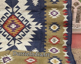 May include: A rectangular rug with a geometric pattern. The rug features a central design in cream, navy blue, and accents of red, yellow, and purple. The border is olive green with repeating geometric shapes. The rug has a fringed edge and the text "DurrieIndiaArt".