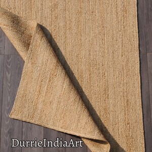 Pode incluir: Um tapete de fibra natural feito de juta, um material sustent&aacute;vel e dur&aacute;vel. O tapete &eacute; de cor castanho claro e tem uma textura tecida. O tapete est&aacute; dobrado ao meio e est&aacute; sobre um piso de madeira. O texto "DurrieIndiaArt" &eacute; vis&iacute;vel no canto inferior esquerdo da imagem.