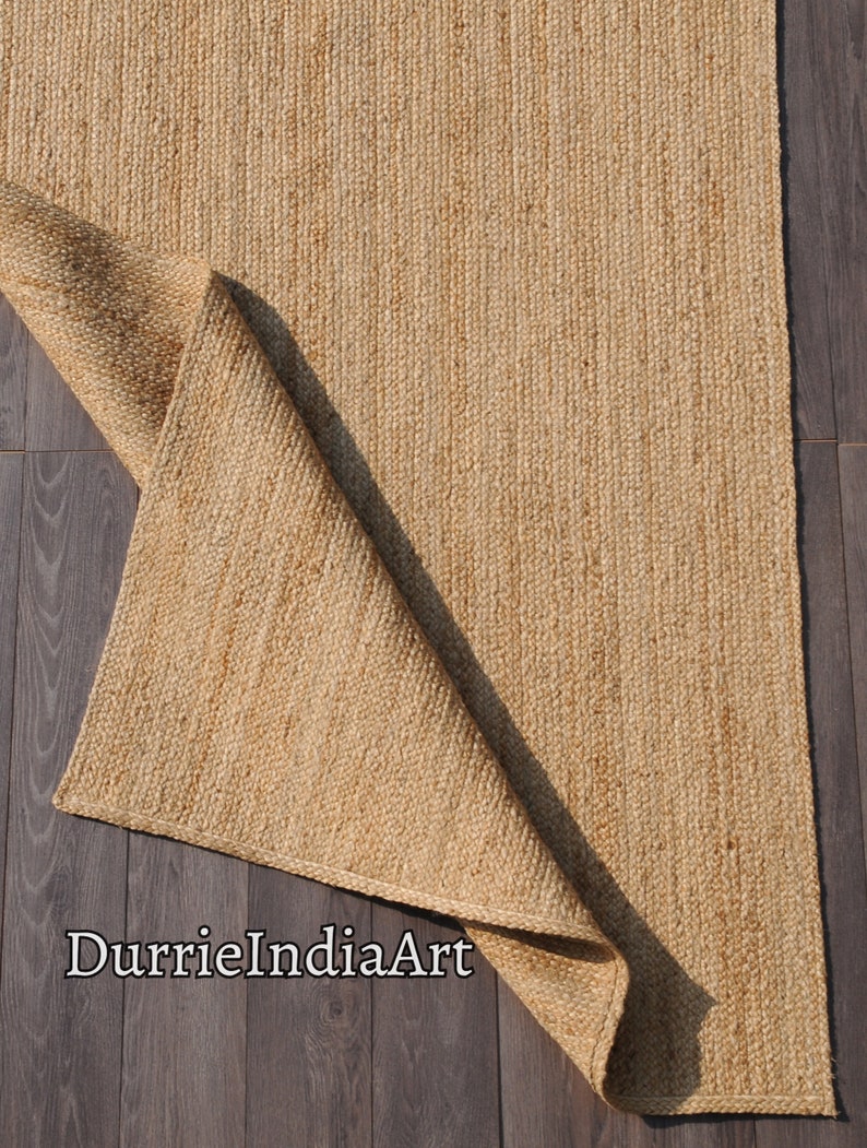 Pode incluir: Um tapete de fibra natural feito de juta, um material sustent&aacute;vel e dur&aacute;vel. O tapete &eacute; de cor castanho claro e tem uma textura tecida. O tapete est&aacute; dobrado ao meio e est&aacute; sobre um piso de madeira. O texto "DurrieIndiaArt" &eacute; vis&iacute;vel no canto inferior esquerdo da imagem.