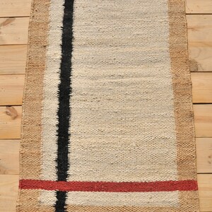 May include: A rectangular jute rug with a white center and a black stripe down the center. The rug has a tan border and a red stripe across the bottom.