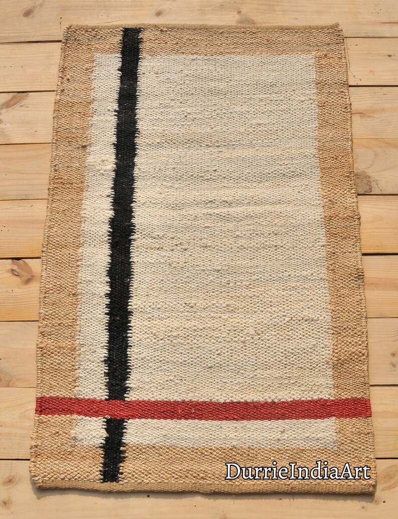 May include: A rectangular jute rug with a white center and a black stripe down the center. The rug has a tan border and a red stripe across the bottom.