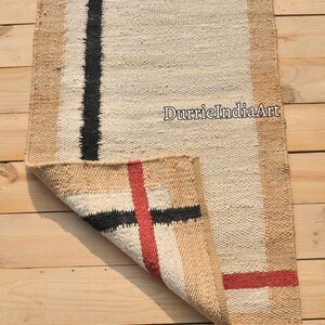 May include: A rectangular jute rug with a white center and a brown border. The rug has black and red stripes woven into the design. The rug is folded in half and the text "DurrieIndiaArt" is visible in the center of the rug.