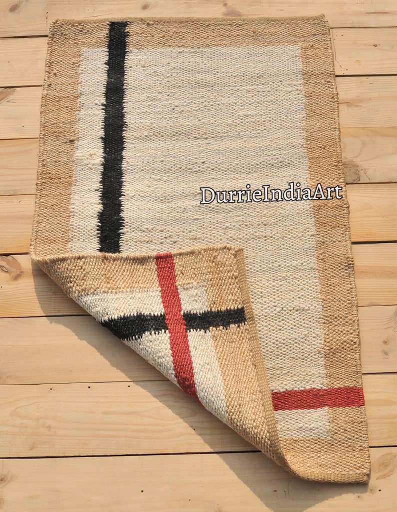 May include: A rectangular jute rug with a white center and a brown border. The rug has black and red stripes woven into the design. The rug is folded in half and the text "DurrieIndiaArt" is visible in the center of the rug.