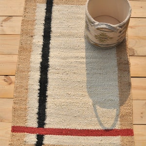 May include: A handwoven rug with a beige background, black stripes, and a red stripe. The rug is made of natural fibers and has a rustic look. The rug is shown with a woven basket.
