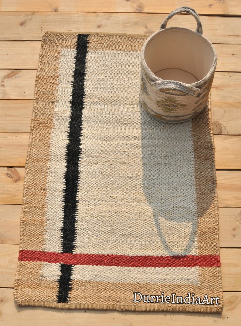 May include: A handwoven rug with a beige background, black stripes, and a red stripe. The rug is made of natural fibers and has a rustic look. The rug is shown with a woven basket.