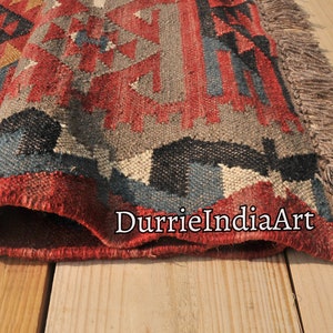 May include: Close-up of a red, blue, and gray patterned rug with fringe. The rug is folded in half and the fringe is visible on the right side of the image. The rug is on a wooden surface.