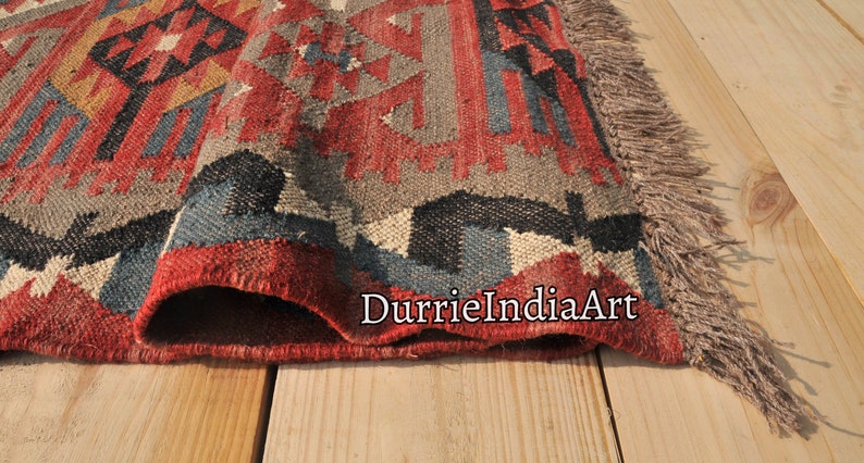 May include: Close-up of a red, blue, and gray patterned rug with fringe. The rug is folded in half and the fringe is visible on the right side of the image. The rug is on a wooden surface.