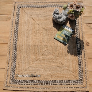 Puede incluir: Una alfombra de fibra natural con un dise&ntilde;o tejido y un borde negro y blanco. La alfombra est&aacute; hecha de yute y tiene un color natural y terroso. La alfombra est&aacute; colocada sobre un suelo de madera.