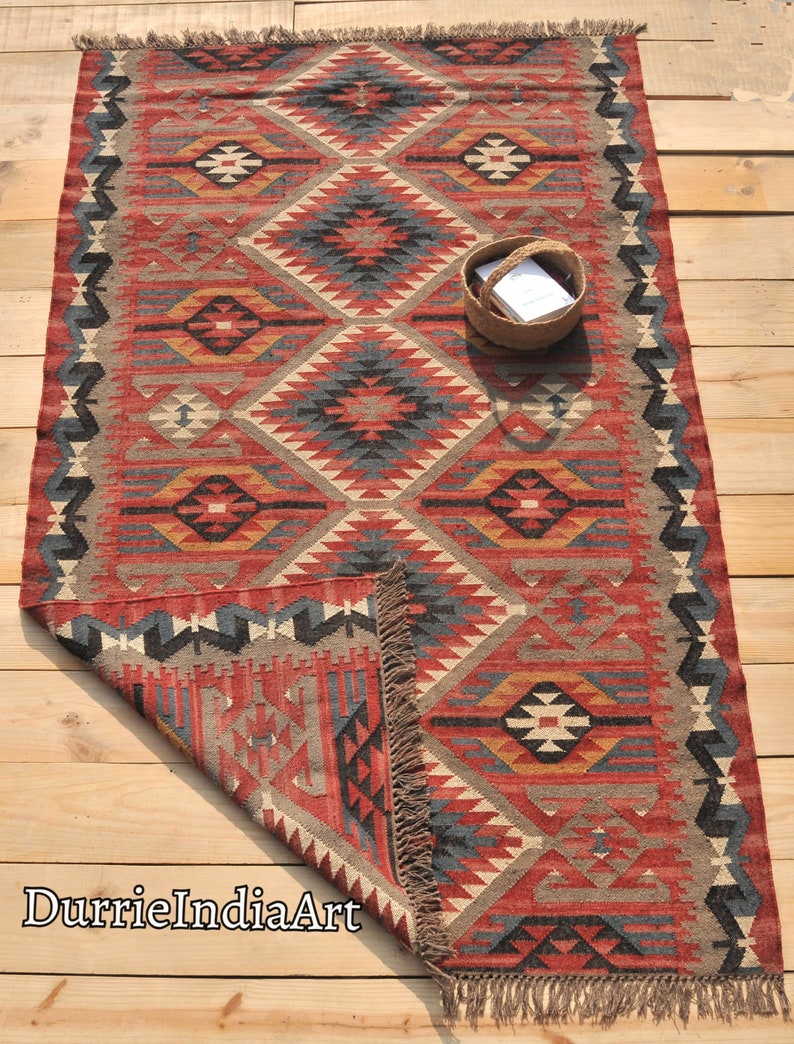 May include: A red, blue, and beige handwoven rug with a geometric pattern. The rug has fringe on all sides and is folded in half. The rug is on a wooden floor.