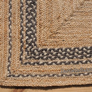 Könnte beinhalten: Ein Naturfaser-Teppich mit gewebtem Design. Der Teppich hat einen Rand aus schwarz-weiß geflochtenem Seil. Der Teppich ist aus Jute und hat eine natürliche, erdige Farbe.